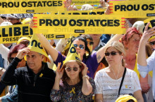 Pla mig de diversos assistents a la manifestació "Us volem a casa" protegint-se del sol i exhibint un rètol on s'hi llegeix "Prou ostatges"
