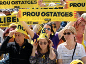 Pla mig de diversos assistents a la manifestació "Us volem a casa" protegint-se del sol i exhibint un rètol on s'hi llegeix "Prou ostatges"